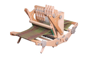 The Ashford Katie, an 8-shaft table loom from Ashford Handicrafts Limited, features a portable wooden frame with green and brown yarn threads. This loom includes several levers, knobs for adjustments, and multiple heddles. The partially completed fabric extends from the front.