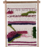 A handwoven tapestry hung on an Ashford Weaving Frame/Tapestry Style Loom by Ashford Handicrafts Limited. The tapestry features a mix of textures and colors including white, pink, purple, green, and dark blue yarns arranged in horizontal bands and various patterns.