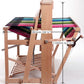 The image shows an Ashford 38" Jack Floor Loom Eight Shaft, 97 cm (38") by Ashford Handicrafts Limited with colorful fabric partially woven on it. There are measurements indicated: width 700mm (27 ½”), depth 985mm (38 ¾”), and height 1100mm (43¼”). The loom has various components and levers visible.