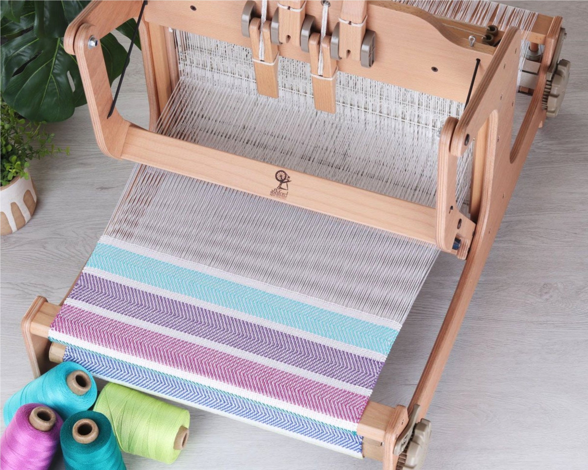 Displayed is the Ashford Brooklyn 16" Table Loom 4-Shaft by Ashford Handicrafts Limited, showcasing a colorful, partially woven fabric in shades of purple, blue, and white. On the floor beside the loom are three spools of thread in turquoise, teal, and pale green. A potted plant in the background completes the scene of this multi-shaft weaving project.