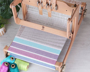 Displayed is the Ashford Brooklyn 16" Table Loom 4-Shaft by Ashford Handicrafts Limited, showcasing a colorful, partially woven fabric in shades of purple, blue, and white. On the floor beside the loom are three spools of thread in turquoise, teal, and pale green. A potted plant in the background completes the scene of this multi-shaft weaving project.