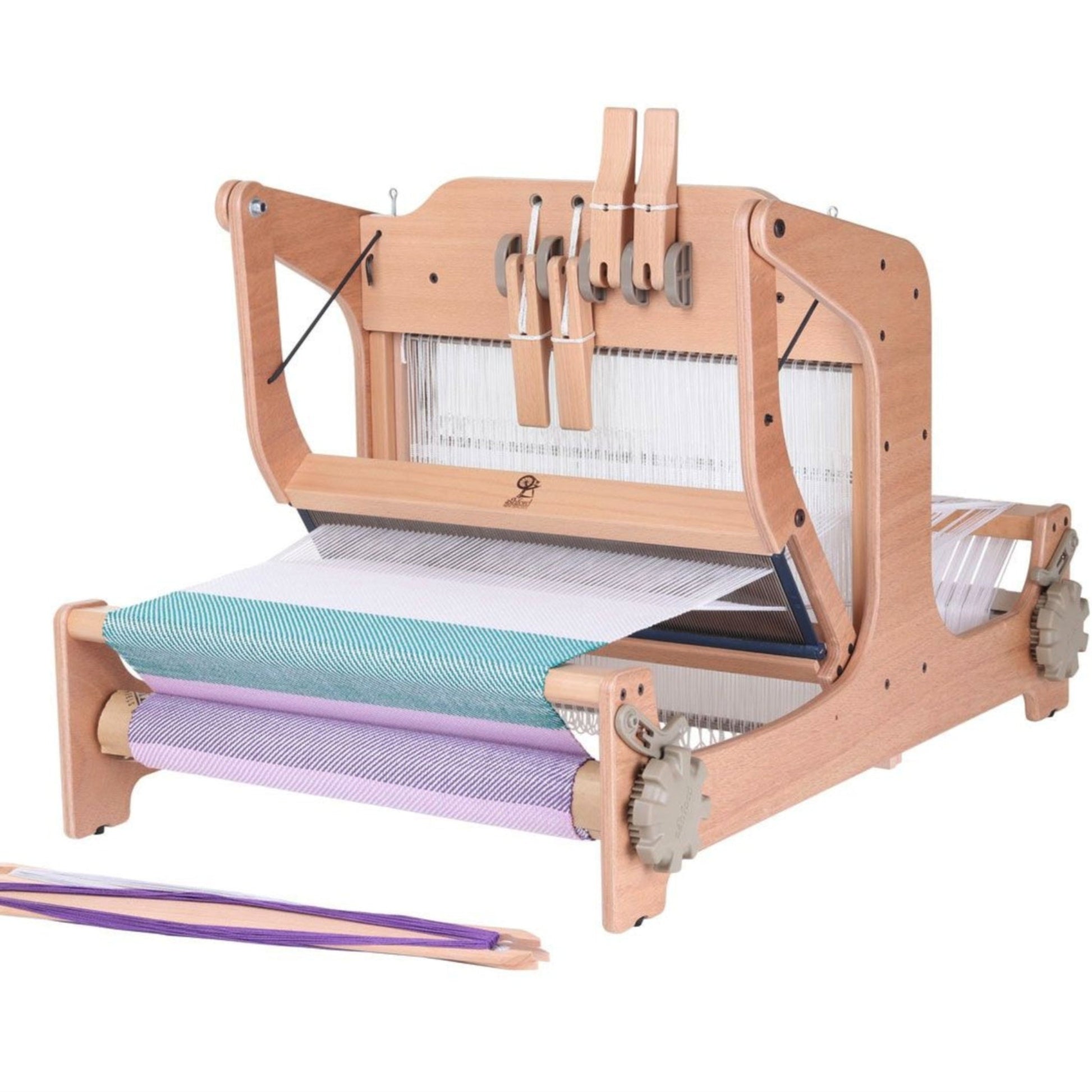 An image of the Ashford Brooklyn 16" Table Loom 4-Shaft by Ashford Handicrafts Limited, showcasing a wooden tabletop weaving loom with threads set up for weaving. The loom features a heddle, beater, and tension knobs, with purple and green yarn wound on the front and back beams. In front of the loom are two wooden shuttles with yarn, making it ideal for multi-shaft weaving projects.