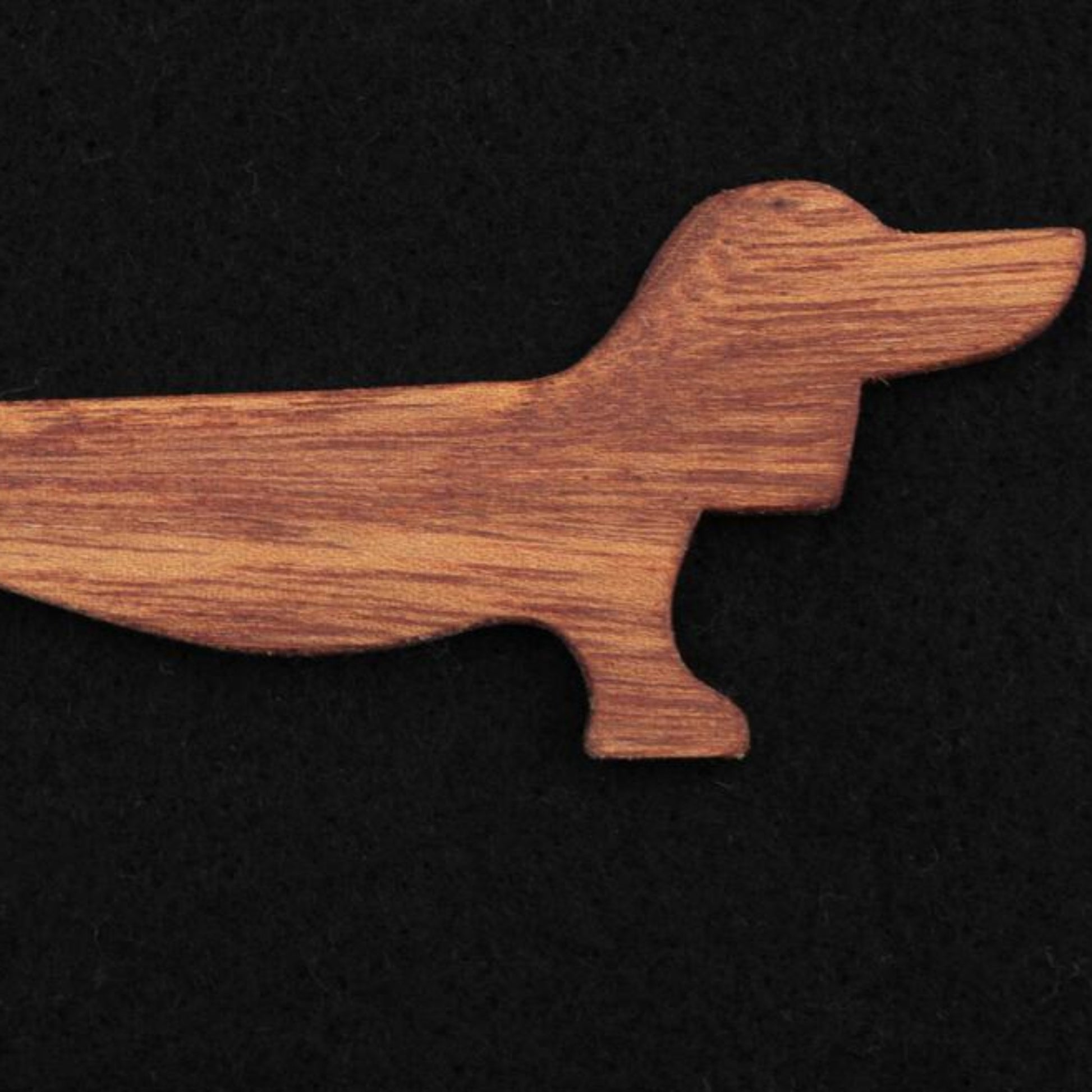 The Dachshund Wiener Dog Weaving Shuttle by Weiner Dog Ranch features a wooden cutout in the shape of a dachshund on a black background. This stylized cutout depicts the dog with its characteristic long body, short legs, and elongated head with a pointed nose. The visible wood grain of the mahogany accessory adds texture to its simple silhouette.