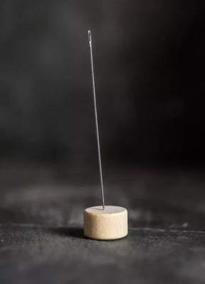 A sewing needle from the Merchant & Mills Long Darners collection stands upright, embedded in a circular wooden base, set against a dark, blurred background. The needle's eye is visible at the top, creating a minimalist and focused composition.