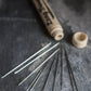 A close-up shot of various Merchant & Mills Long Darners fanned out next to an open, beige cylindrical container on a dark, textured surface. The container has a fitted cap lying beside it and the text "Merchant & Mills" is partially visible on the side.