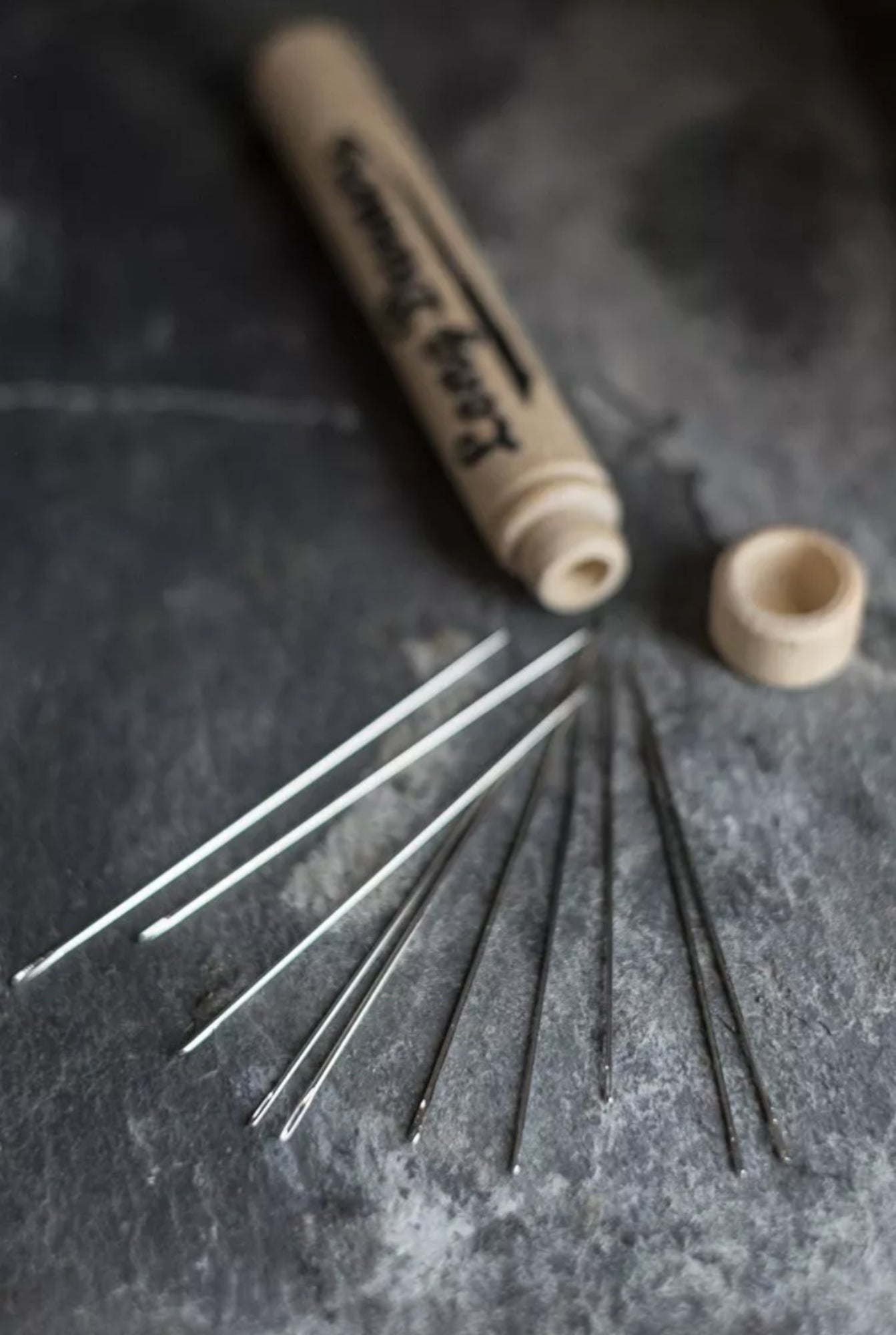 A close-up shot of various Merchant & Mills Long Darners fanned out next to an open, beige cylindrical container on a dark, textured surface. The container has a fitted cap lying beside it and the text "Merchant & Mills" is partially visible on the side.