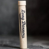 A wooden Merchant & Mills Long Darners holder is standing upright on a dark surface. The holder is cylindrical and features the inscription "Merchant & Mills Long Darners" in a stylish, cursive font along its side. The background is slightly blurred to emphasize the holder.