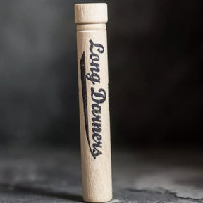 A wooden Merchant & Mills Long Darners holder is standing upright on a dark surface. The holder is cylindrical and features the inscription "Merchant & Mills Long Darners" in a stylish, cursive font along its side. The background is slightly blurred to emphasize the holder.