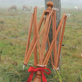 A hand-crafted Maine Made Cherry Swift from Thomas Siske stands against a fencepost in a misty field. A spider web is visible between the swift and the post. Sheep graze in the blurred background. A red ribbon is tied around the base of this superior quality, Shaker-styled swift.