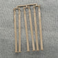 A set of six beige plastic stakes featuring arrow-shaped tops and flat bases, linked by a slender frame, resembling Ashford Handicrafts Limited accessories, is laid out on a textured gray fabric background.