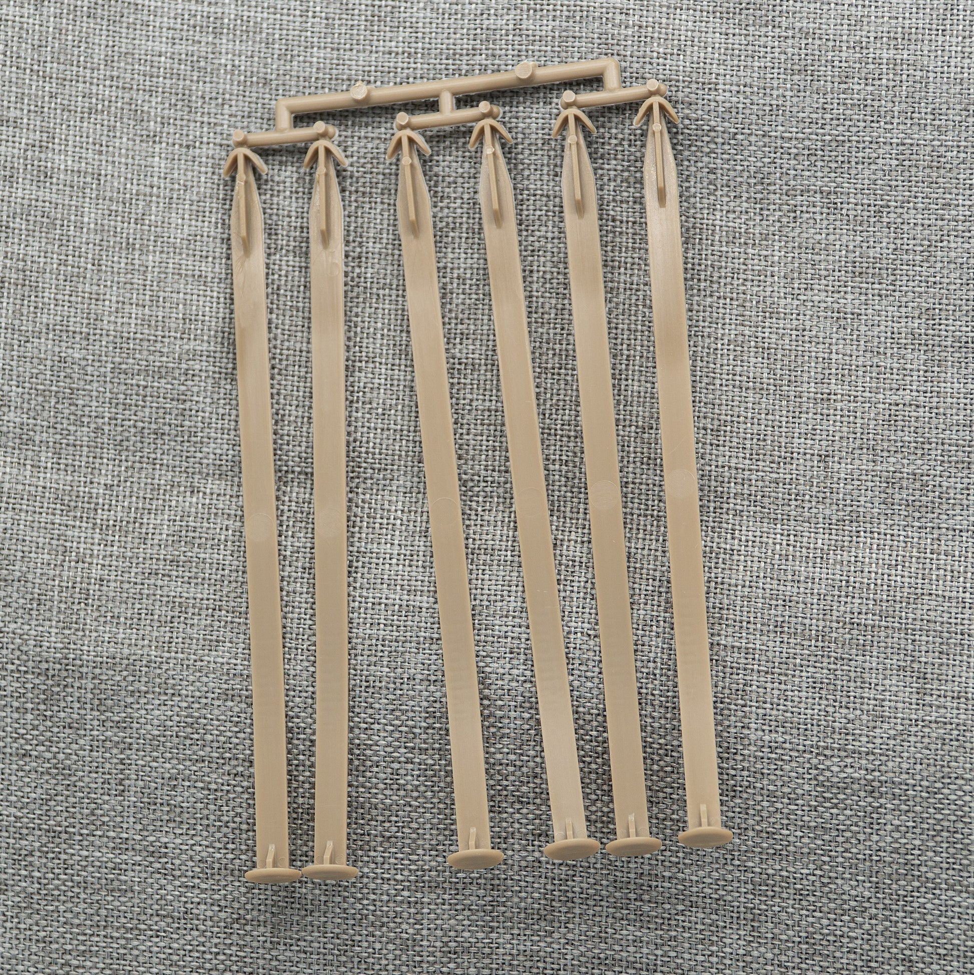 A set of six beige plastic stakes featuring arrow-shaped tops and flat bases, linked by a slender frame, resembling Ashford Handicrafts Limited accessories, is laid out on a textured gray fabric background.