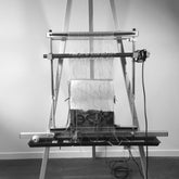 A black and white photograph depicts a weaving loom with partially completed fabric on it, mounted on an adjustable stand. Threads are neatly aligned, and a pattern emerges in the woven section. The loom is supported by what appears to be the Mirrix Tripod Tapestry Loom Floor Stand from Mirrix Tapestry Looms. This 5.5 ft floor stand is placed against a plain wall surrounded by cables and various weaving tools.