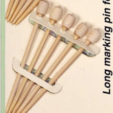 A close-up of a pack of Clover Bamboo Marking Pins by Accessories Unlimited, ideal for thick knitting fabrics. The pins feature wooden heads and are secured in a cardboard holder. Some additional loose pins are partially visible in the bottom left corner of the image.