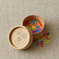 A round cardboard container labeled "Colored Ring Stitch Markers" by Cocoknits is open, revealing numerous small circular knitting accessories in various bright colors such as red, green, blue, yellow, orange, and purple. Some markers are scattered on the beige fabric background.