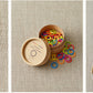 Three images showcasing vibrant, rainbow-colored Cocoknits Colored Ring Stitch Markers and their application. Left: Seven stitch markers in a vertical line, arranged by color. Center: A container with nylon-coated steel Cocoknits Colored Ring Stitch Markers spilling out. Right: A knitting project with these essential knitting accessories placed along the needle.