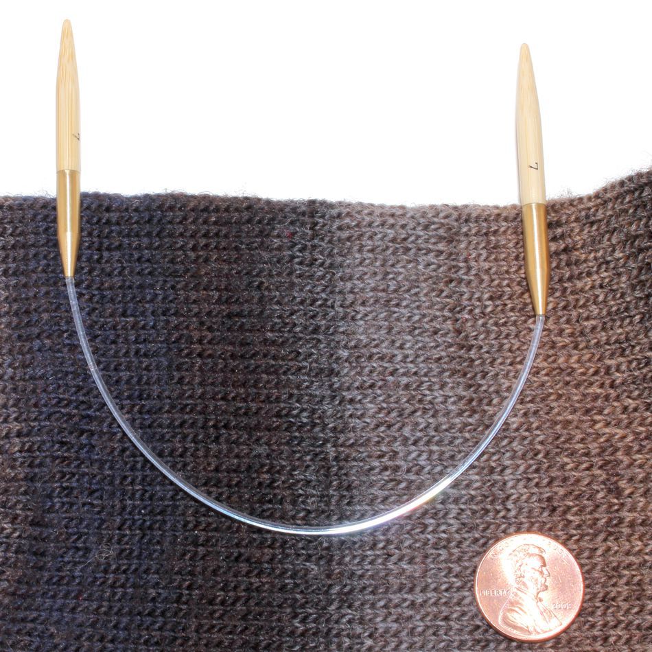 A circular bamboo knitting needle with wooden tips and a flexible transparent cord from Accessories Unlimited lies against a dark gray and brown knitted fabric. The Circular Bamboo Knitting Needles feature a flexible rotating joint for ease of use, with a penny placed next to the fabric to showcase the relative size.