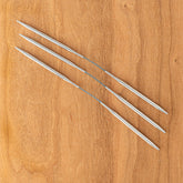 Four Addi FlexiFlips Circular Needles from Skacel, made of silver-colored metal, are arranged diagonally on a wooden surface. These flexible double-pointed needles have a slight curve in the middle and are tapered to a point at each end.