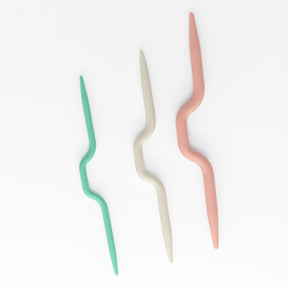 Three Bent Cable Stitch Holders from Accessories Unlimited, in green, beige, and peach colors, are laid out in a vertical, slightly diagonal arrangement on a white background. Resembling US size 8 cable needles, each holder features a wavy, zigzag design in the middle.