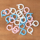 A variety of Clover Split Ring Stitch Markers from Bryson Distributing, Inc., featuring pink, blue, and white plastic markers shaped like hooks, are essential knitting accessories scattered on a wooden surface.