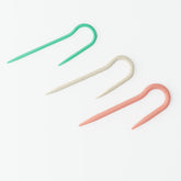 Three J-Cable Stitch Holders by Bryson Distributing, Inc. are arranged on a white background. They are curved into a hook shape and come in three colors: green, cream, and coral. Designed to fit needles up to size 8, they also work perfectly with cable needles of different sizes.