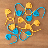 A collection of Accessories Unlimited Locking Ring Stitch Markers in orange and blue, made from plastic and perfect for bulky yarns, is spread across a wooden surface. These jumbo markers feature a curved shape with a small latch to secure yarn or threads, making them ideal for knitting and crochet projects.