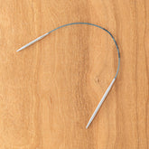 A pair of Skacel Addi EasyKnit Turbo Circular Needles, featuring a blue cord connecting two 10" silver metal needle tips, arranged in a semi-circular shape on a wooden surface.