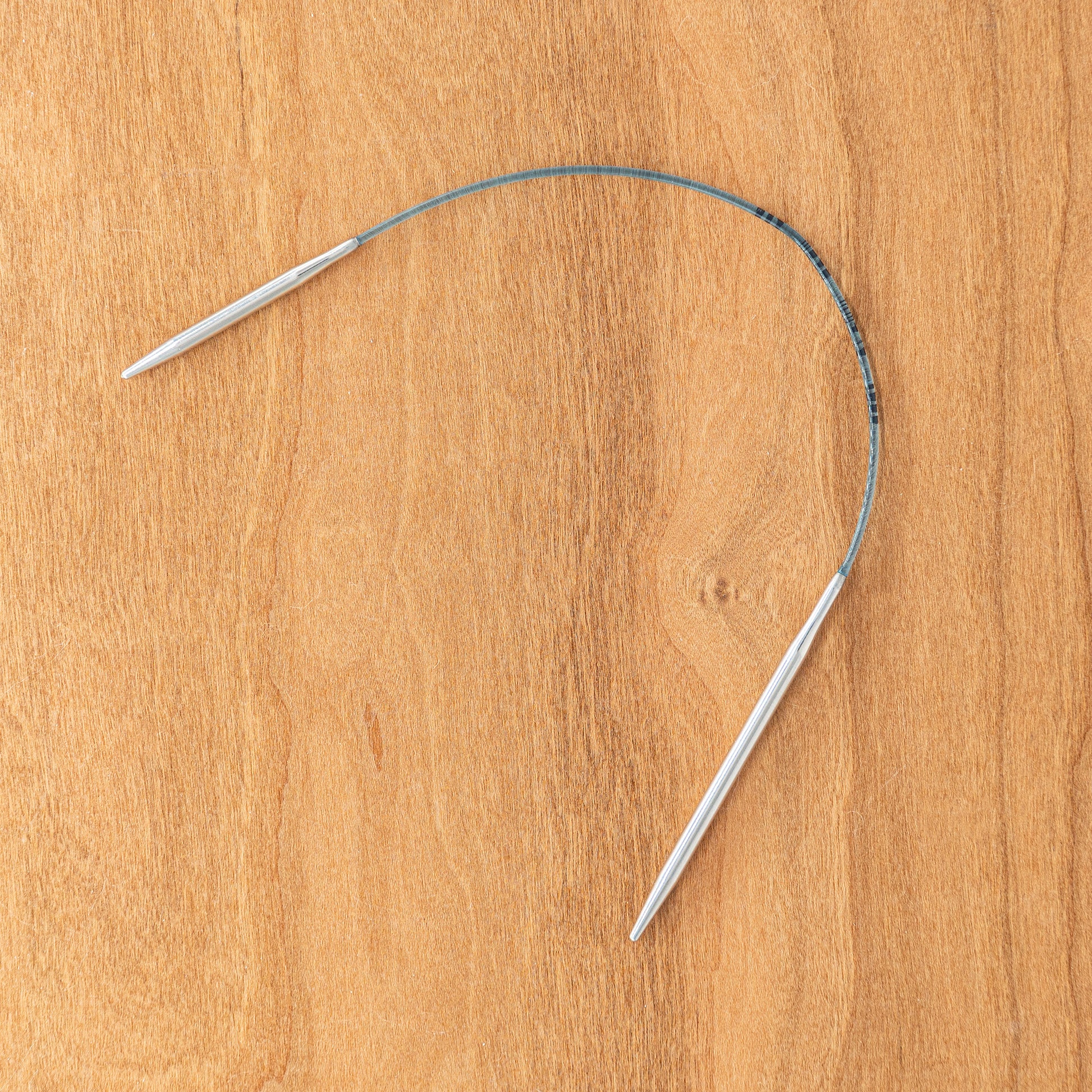 A pair of Skacel Addi EasyKnit Turbo Circular Needles, featuring a blue cord connecting two 10" silver metal needle tips, arranged in a semi-circular shape on a wooden surface.