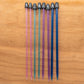 On a light wooden surface, a row of eight colorful Crystalite Straight Knitting Needle Sets from Accessories Unlimited, each 10 inches long, is displayed. Perfect for small projects like knitting scarves, the vibrant plastic needles are organized by size from left to right. The colors graduate from pink to blue and each needle is topped with black caps that display their sizes in both metric and US units.