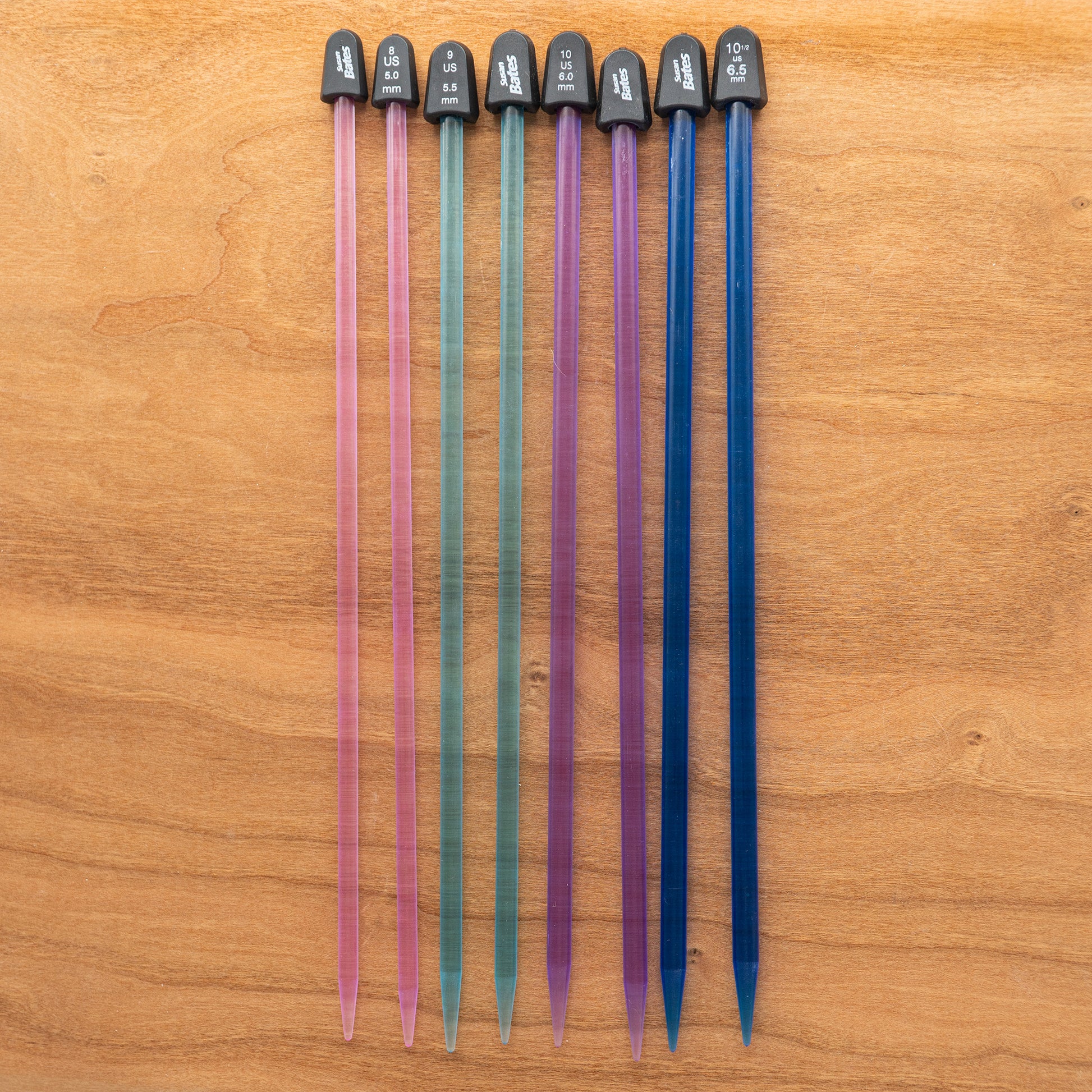 On a light wooden surface, a row of eight colorful Crystalite Straight Knitting Needle Sets from Accessories Unlimited, each 10 inches long, is displayed. Perfect for small projects like knitting scarves, the vibrant plastic needles are organized by size from left to right. The colors graduate from pink to blue and each needle is topped with black caps that display their sizes in both metric and US units.