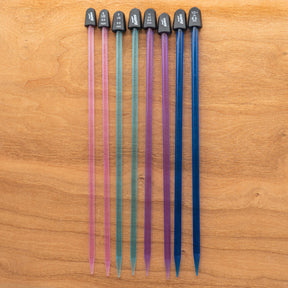 On a light wooden surface, a row of eight colorful Crystalite Straight Knitting Needle Sets from Accessories Unlimited, each 10 inches long, is displayed. Perfect for small projects like knitting scarves, the vibrant plastic needles are organized by size from left to right. The colors graduate from pink to blue and each needle is topped with black caps that display their sizes in both metric and US units.