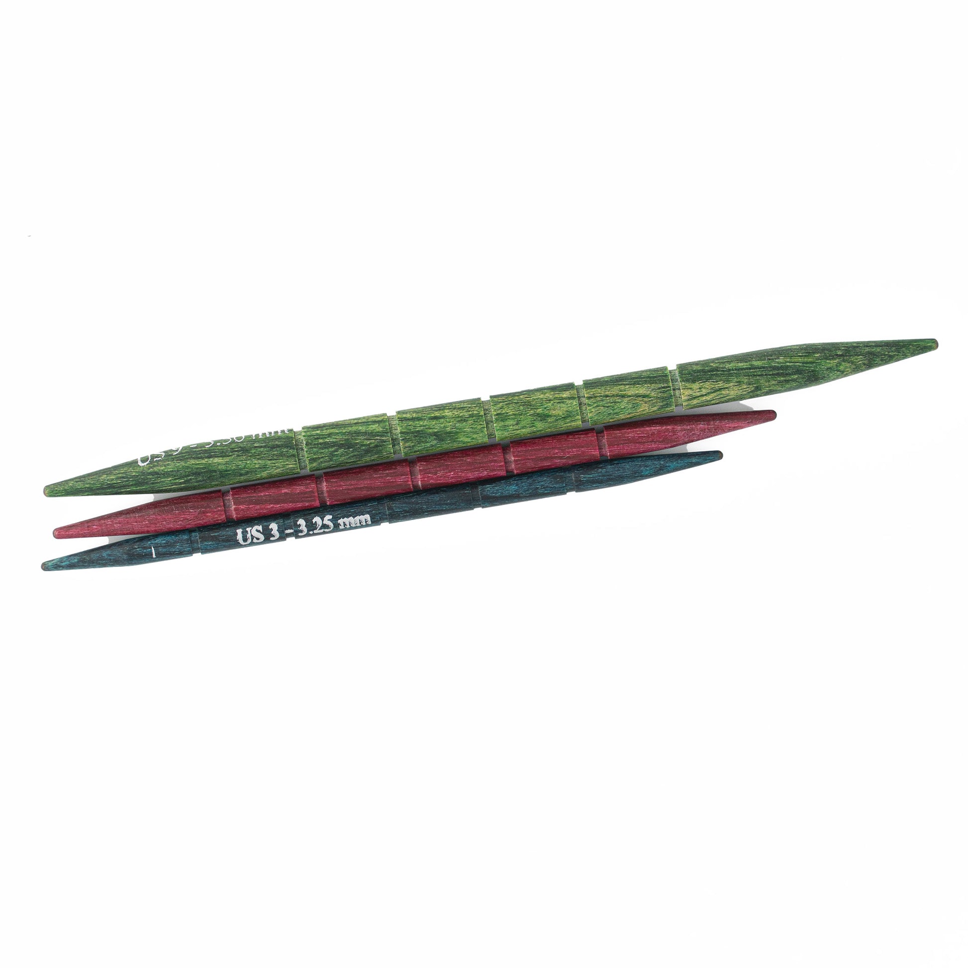 Three horizontal double-pointed knitting needles, one green, one red, and one blue. The blue needle is inscribed with "US 3 - 3.25 mm." Each laminated birch needle features evenly spaced grooves. These cable needles are from the Dreamz Cable Needles collection by Knitter's Pride, available through Accessories Unlimited.