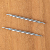 Two silver Nova Platina Interchangeable Knitting Needles from Accessories Unlimited, each with a 4.5" tip, are positioned horizontally on a wooden surface. The needles have a smooth, shiny finish and gradually taper to a point at each end. The wood has a light, natural grain pattern.