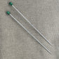 Two long, thin Nova Platina Single Point Knitting Needles from Accessories Unlimited, similar to straight needles, are placed parallel on a textured, light-colored fabric surface. Each needle features a green rectangular handle at one end and a sharp point at the other.