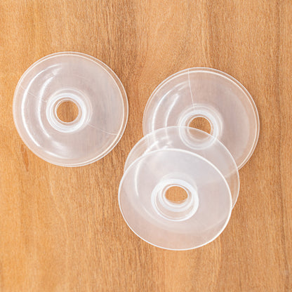 Three EZ Bobs Flip-top Intarsia Bobbins from Bryson Distributing, Inc. with central holes are spread out on a wooden surface. The bobbins appear to be identical and translucent, showing a slight reflection of light. They are slightly overlapping each other.