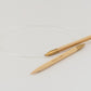 Two knitting needles connected by a transparent cord lie on a white surface. The handles of the Accessories Unlimited Circular Bamboo Knitting Needles are a light wood color with golden tips. One is marked with the size "40" in small text, and they feature a flexible rotating joint for smooth knitting.