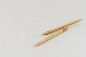 Two knitting needles connected by a transparent cord lie on a white surface. The handles of the Accessories Unlimited Circular Bamboo Knitting Needles are a light wood color with golden tips. One is marked with the size "40" in small text, and they feature a flexible rotating joint for smooth knitting.