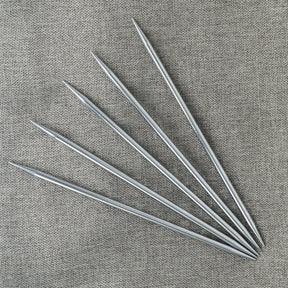 Five Nova Platina Double-Point Knitting Needles from Accessories Unlimited are neatly arranged in a fan-like pattern on a textured, gray fabric background. The high-quality chrome finishing highlights their smooth, shiny surfaces against the fabric.