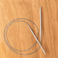 A pair of Skacel's Addi Turbo Circular Knitting Needles with a flexible blue cable are laid out on a wooden surface. The cable forms a loop, and the US sizes 0-8 needles are positioned diagonally across each other. Ideal for knitting cuffs, collars, or doll clothes.