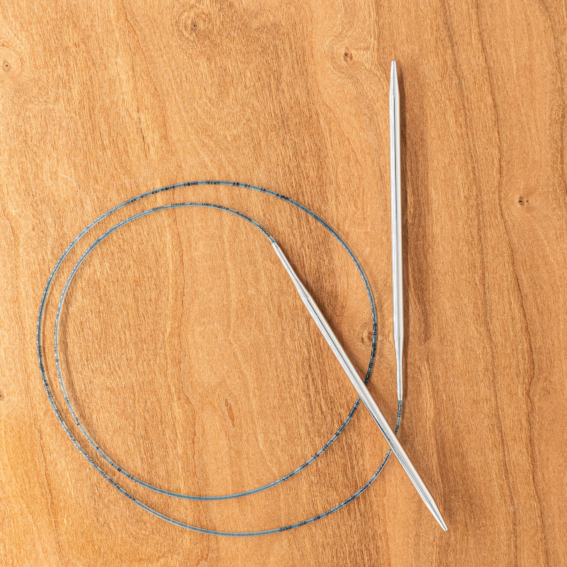 A pair of Skacel's Addi Turbo Circular Knitting Needles with a flexible blue cable are laid out on a wooden surface. The cable forms a loop, and the US sizes 0-8 needles are positioned diagonally across each other. Ideal for knitting cuffs, collars, or doll clothes.