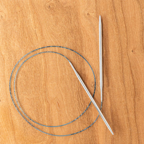 A pair of Skacel's Addi Turbo Circular Knitting Needles with a flexible blue cable are laid out on a wooden surface. The cable forms a loop, and the US sizes 0-8 needles are positioned diagonally across each other. Ideal for knitting cuffs, collars, or doll clothes.