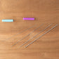 Displayed on a wooden surface are five long sewing needles from the Handy Hands Tatting Needle Sets, neatly arranged in a horizontal row to showcase their varying sizes. Above the needles, two colorful needle threaders—a blue one and a purple one—add a vibrant touch, perfect for working with tatting needles or Pearl Cotton yarn.