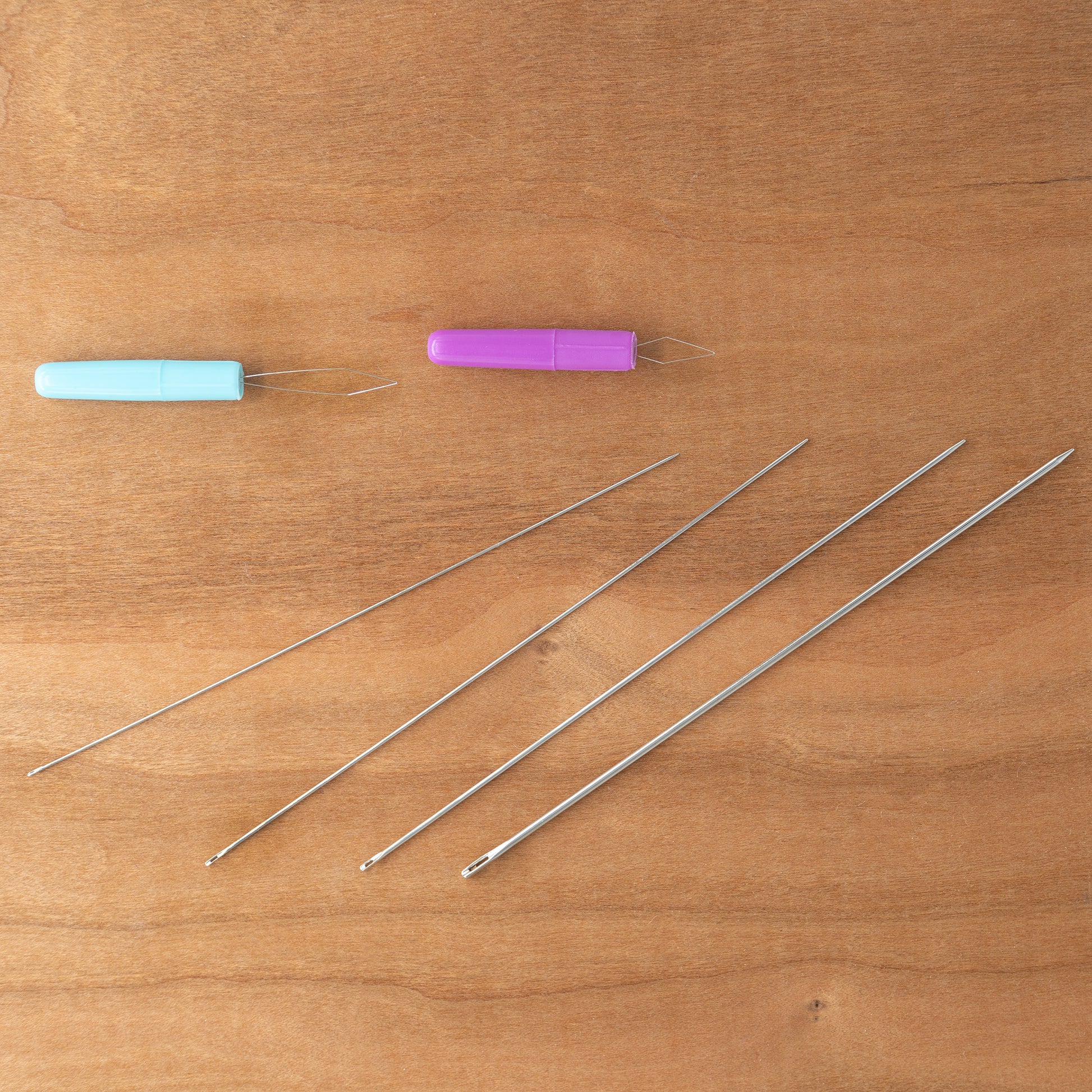 Displayed on a wooden surface are five long sewing needles from the Handy Hands Tatting Needle Sets, neatly arranged in a horizontal row to showcase their varying sizes. Above the needles, two colorful needle threaders—a blue one and a purple one—add a vibrant touch, perfect for working with tatting needles or Pearl Cotton yarn.