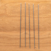 Five Skacel Addi Steel Double Point Knitting Needles, 8", are arranged in a vertical line on a wooden surface. The needles are evenly spaced and parallel to each other, perfect for intricate sock knitting. The wood has a light, natural grain pattern.