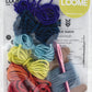 The Loome Kit - Friendship Bracelets from Loome includes an assortment of colorful yarns (purple, blue, light blue, yellow, orange, and red) arranged in loops. The kit also features a pink crochet hook, a brown cardboard spool with blue yarn, and a partially visible instruction sheet. It's perfect for creating vibrant friendship bracelets.