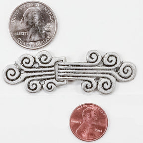 An ornate Antique Silver Metal Clasp from Products from Abroad, featuring intricate swirling designs, is positioned on a white background. For scale reference, a U.S. quarter is placed above and a penny below the embellishment.