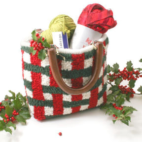 A Basket Weave Purse by Southern Hills Designs, adorned with a red, green, and white plaid pattern and complete with a handle, comes filled with knitting supplies such as Halcyon Rug Wool and needles. The festive theme is accentuated by holly sprigs with red berries arranged around the bag.