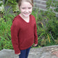 A young child stands outdoors on a rock, smiling at the camera and wearing a red knitted *Wonderful Wallaby For Kids* sweater by *Cottage Creations* and blue pants in kids sizes. They sport sneakers, and there is greenery in the background, including plants and part of a bush. A building with visible windows and siding is behind the child.