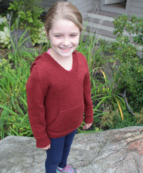 A young child stands outdoors on a rock, smiling at the camera and wearing a red knitted *Wonderful Wallaby For Kids* sweater by *Cottage Creations* and blue pants in kids sizes. They sport sneakers, and there is greenery in the background, including plants and part of a bush. A building with visible windows and siding is behind the child.