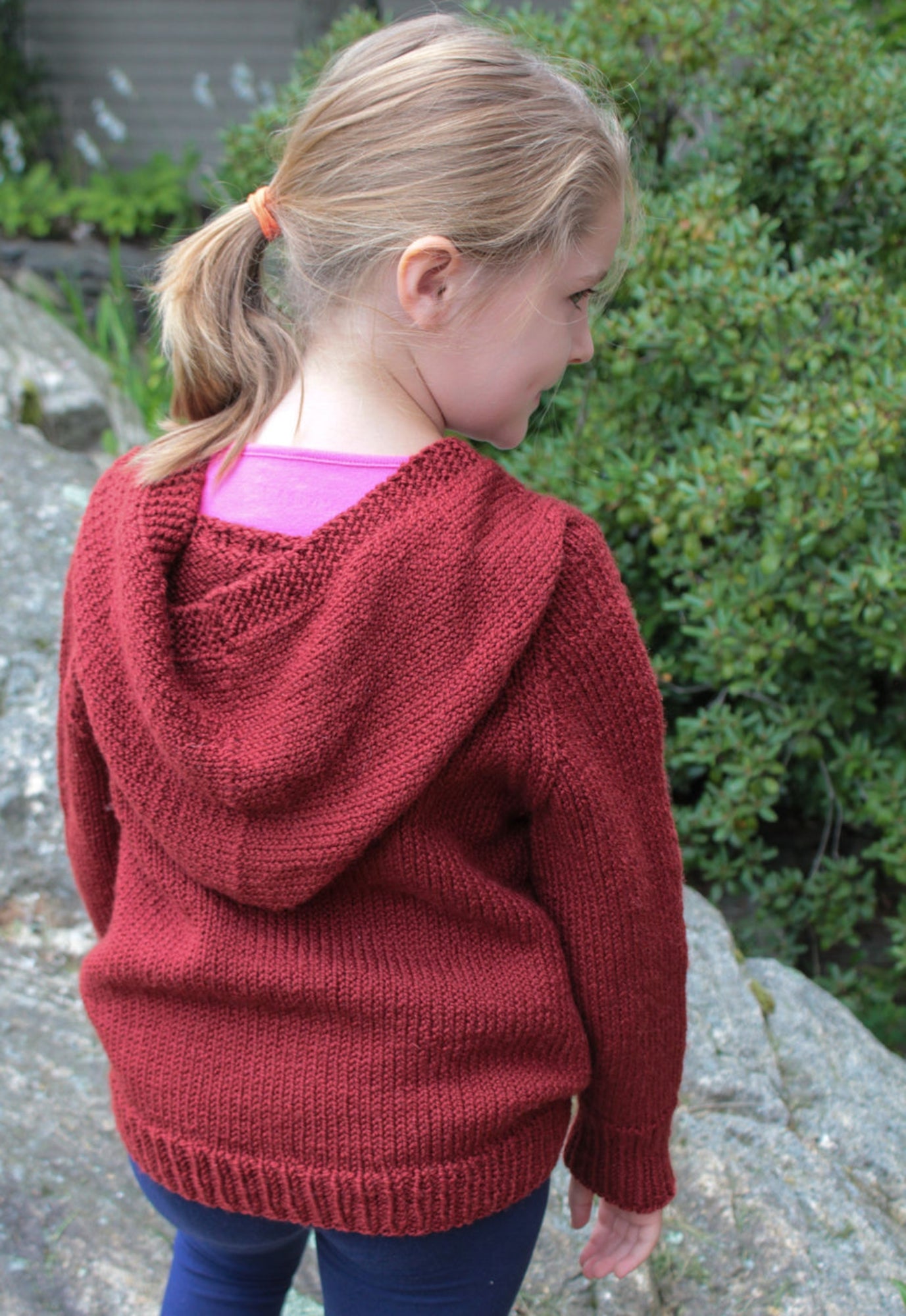 A young girl with blonde hair tied in a ponytail is standing with her back to the camera, wearing Cottage Creations' "A New Generation... Wonderful Wallaby For Kids." The maroon hooded sweatshirt is layered over a pink shirt and paired with blue pants, perfect for kids sizes. She appears to be outside, with green foliage in the background.