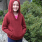 A smiling young girl stands outdoors, wearing a red Cottage Creations Wonderful Wallaby For Grown-Ups knitted hooded sweatshirt with her hands in the front hand-warming pockets. She has blue pants on and is standing in front of greenery and a rock.